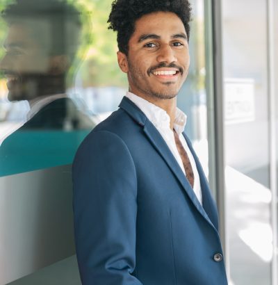 Confident African American male entrepreneur in blazer smiling and looking at camera while standing near reflecting glass wall of modern office building in daylight